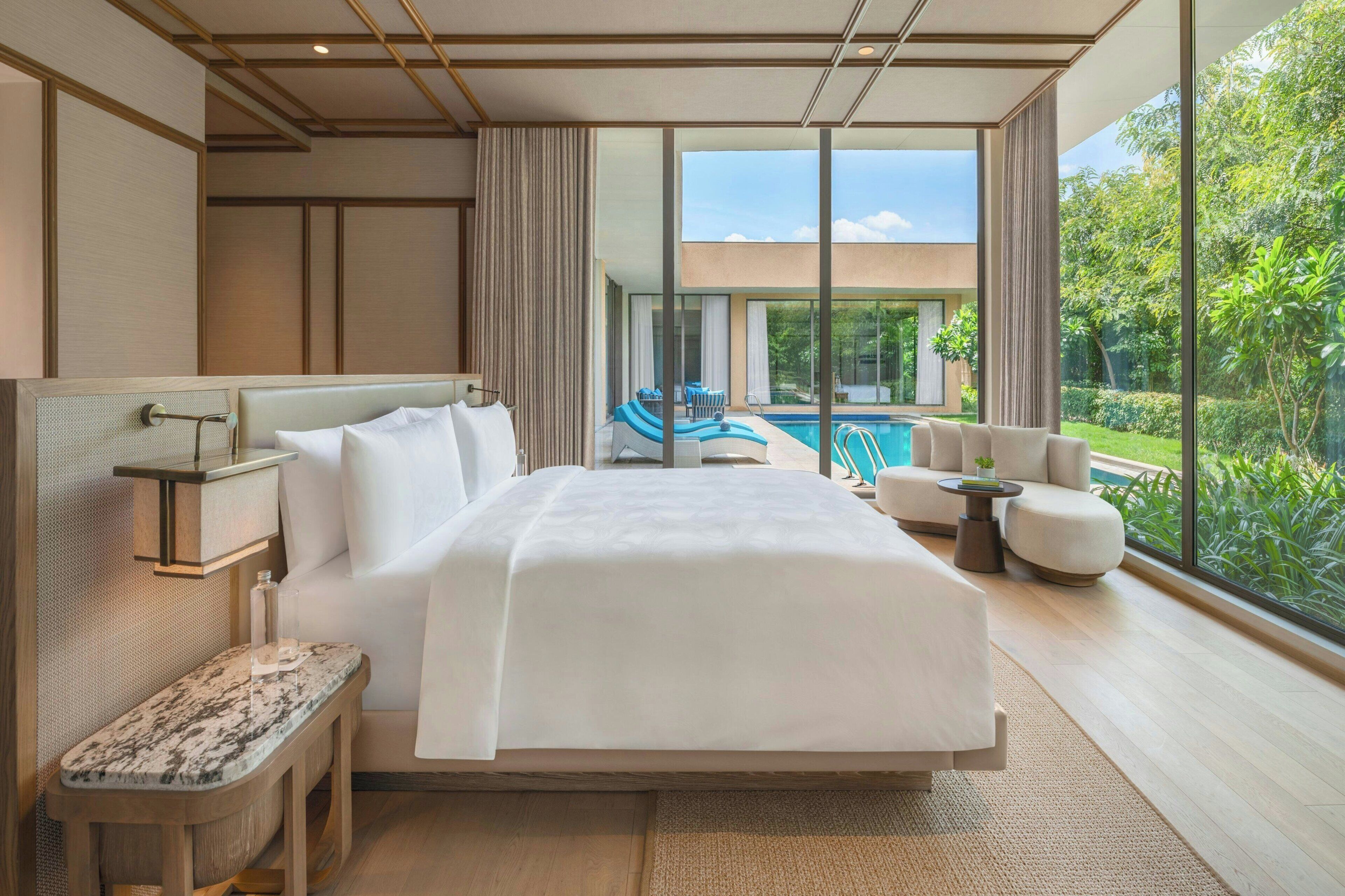 A hotel room with a large bed, a sitting area with a pool view, and a balcony with a view of the greenery.