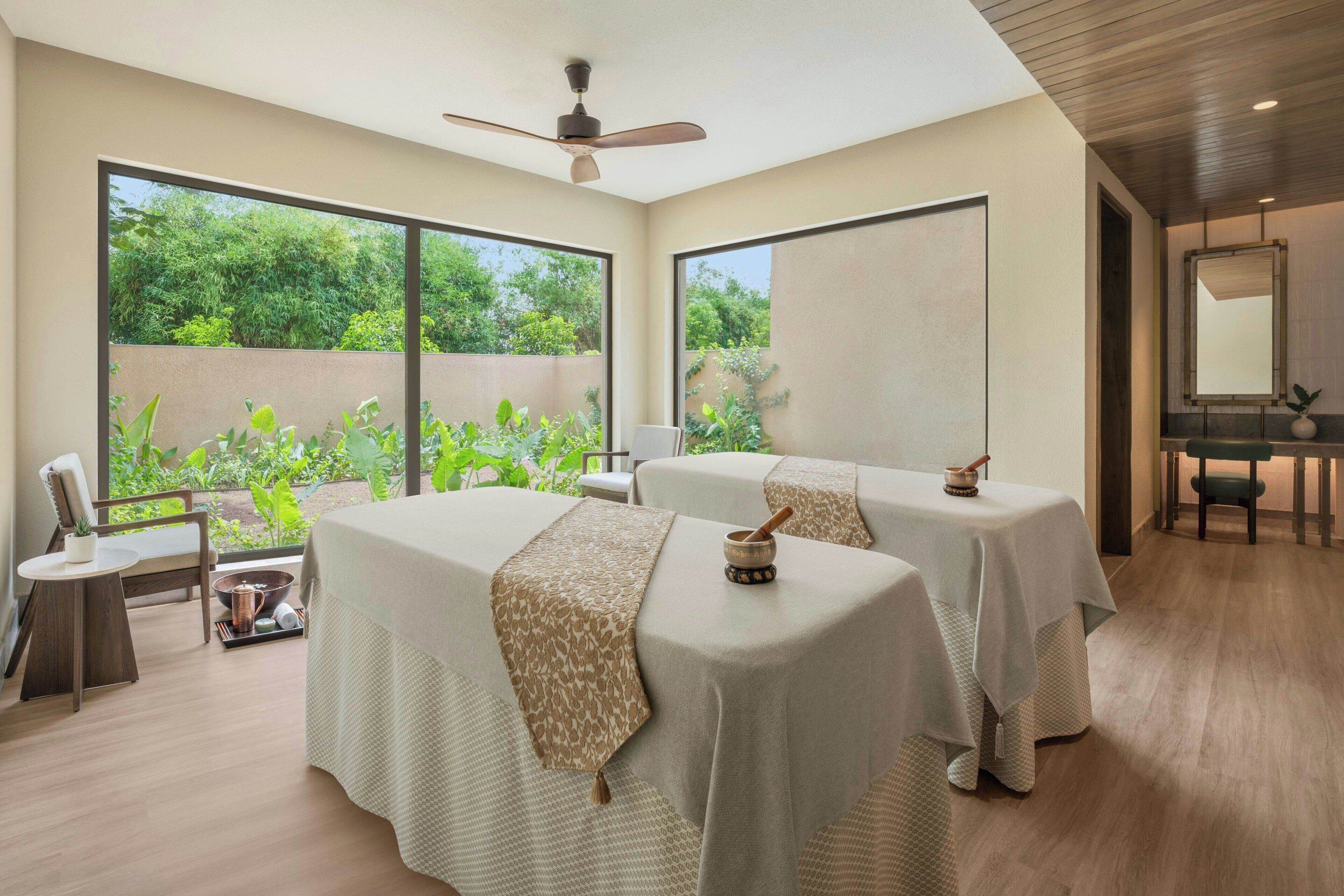 Un cuarto de spa con dos camillas de masaje, un ventilador de techo, ventanas amplias y vistas a la vegetación exterior.