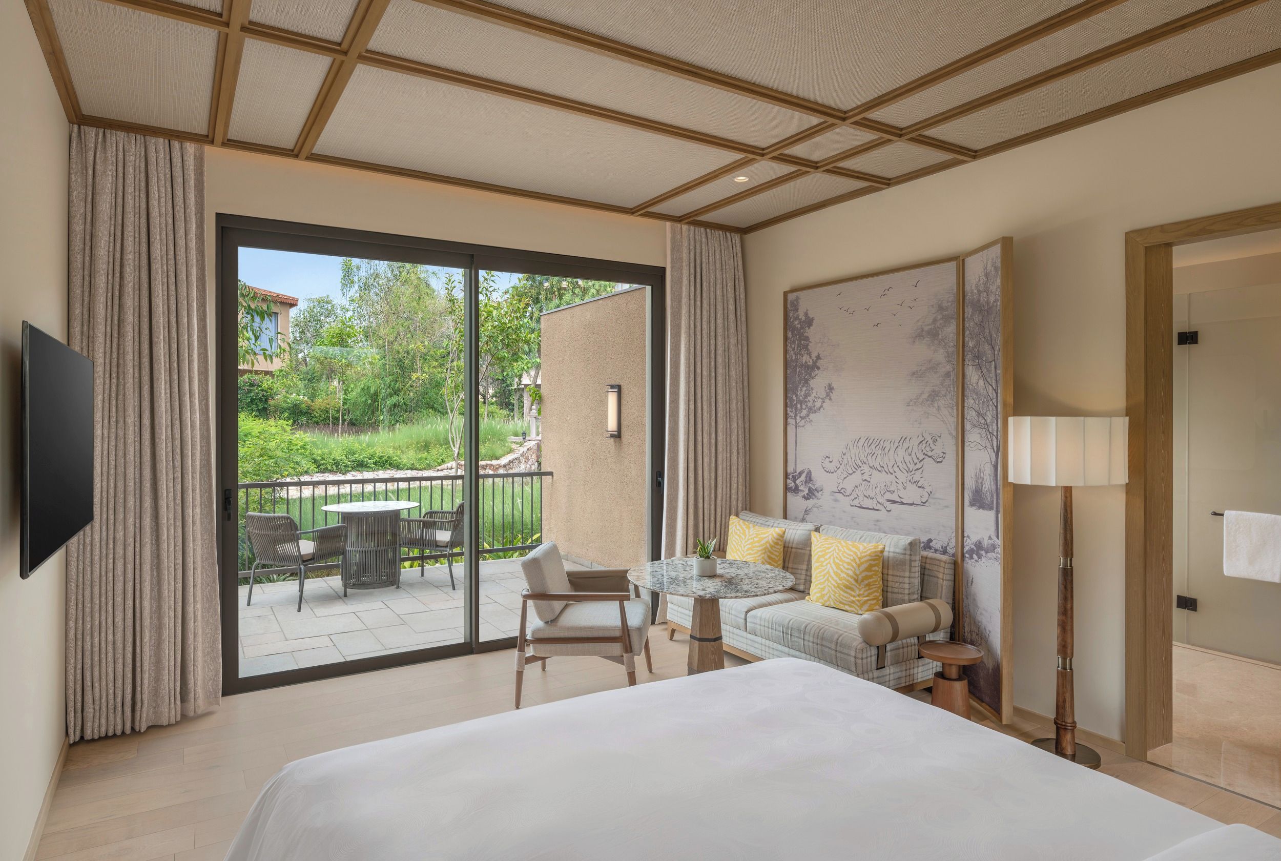 A hotel room with a large bed, a balcony with outdoor furniture, and a view of greenery.