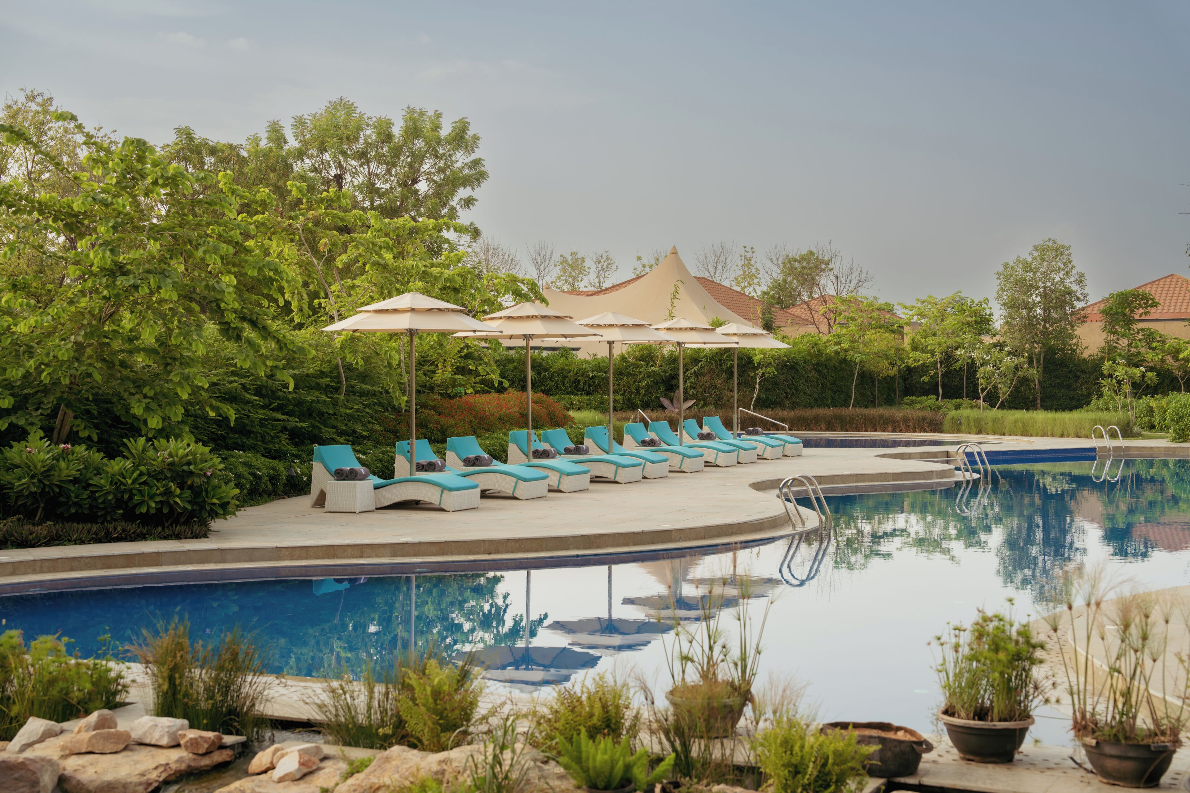 A pool area with lounge chairs and umbrellas, surrounded by greenery and a building in the background.