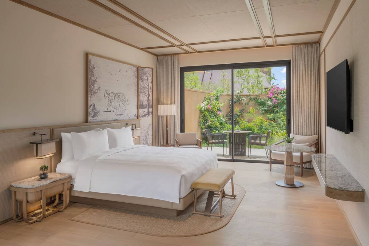 A hotel room with a bed, bedside table, lamp, TV, and a view of a garden through a sliding door.