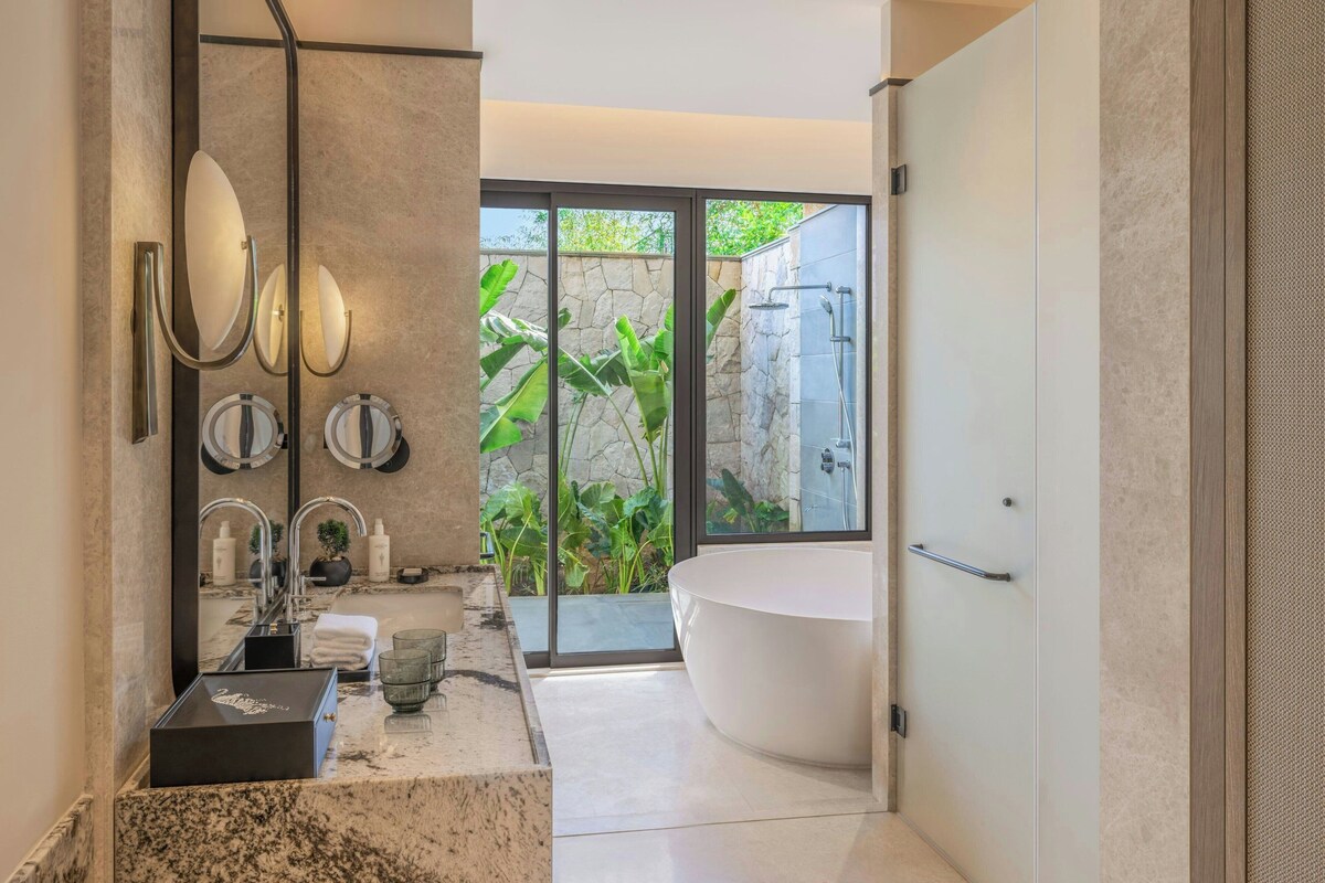 A modern bathroom with a freestanding bathtub, a large mirror, and a view of greenery outside.