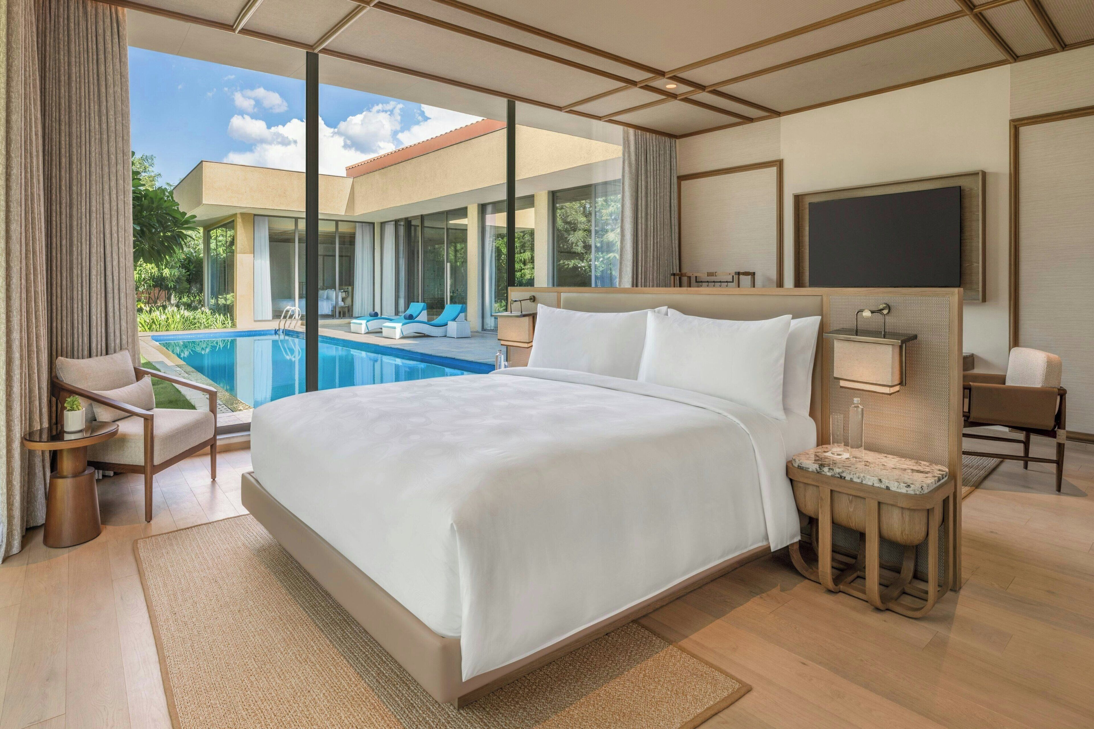 A hotel room with a large bed, a bedside table, a TV, and a view of a swimming pool and outdoor area.
