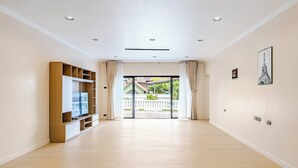 A spacious room with a wooden floor, a built-in TV unit, and a view of a balcony with outdoor scenery.