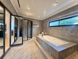 Une salle de bain moderne avec une grande baignoire, une douche en verre et une vue sur les arbres par la fenêtre.
