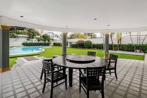 Un patio couvert avec une table ronde et des chaises, une piscine en arrière-plan et une pelouse bien entretenue.