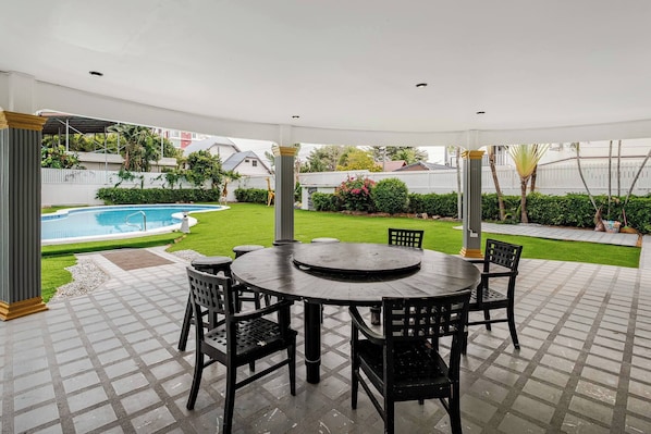Un patio couvert avec une table ronde et des chaises, une piscine en arrière-plan et une pelouse bien entretenue.