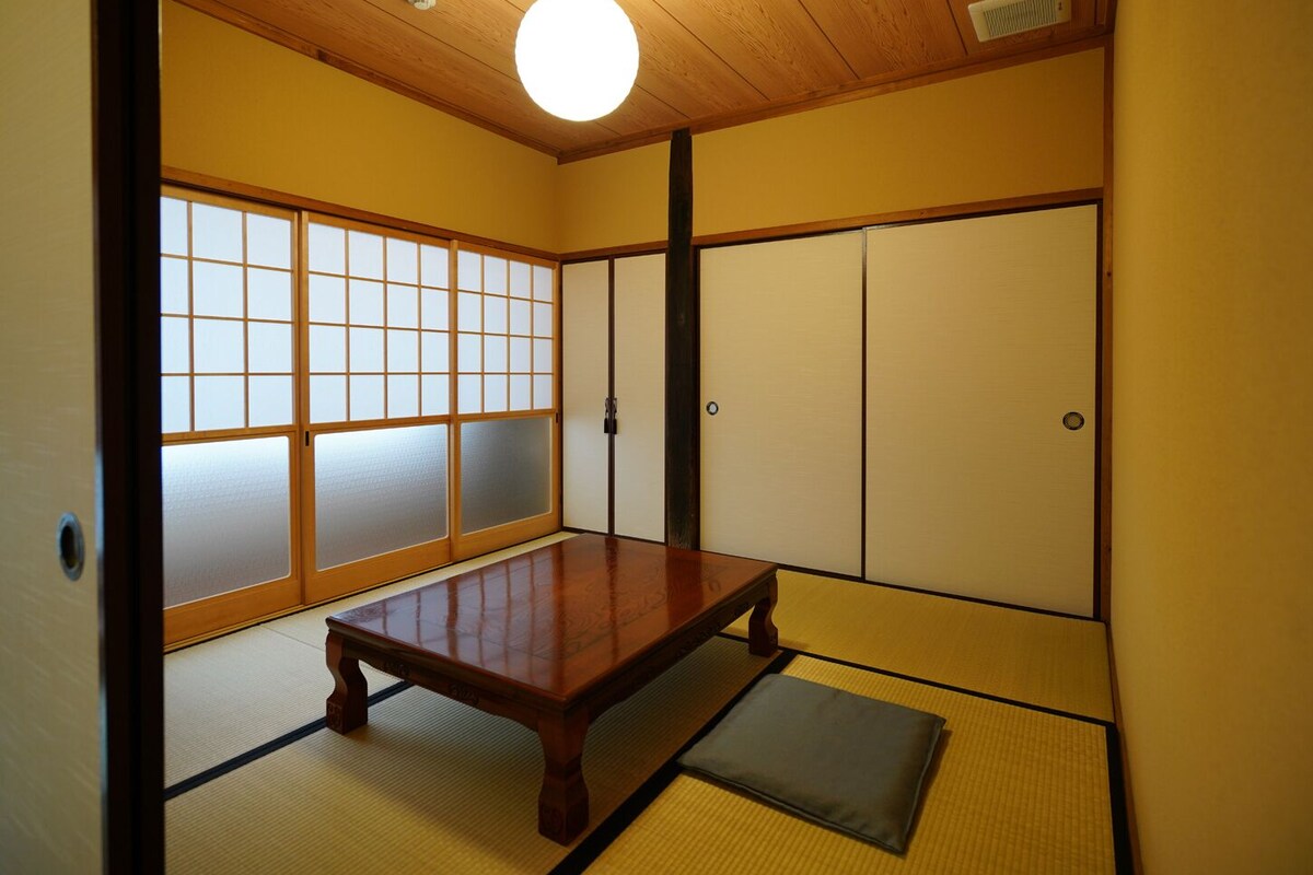 Una habitación japonesa tradicional con suelo de tatami, una mesa de madera baja, puertas correderas y una lámpara en el techo.