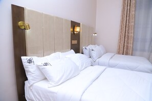 A hotel room with two beds, a wooden headboard, a wall-mounted lamp, and a window with curtains.