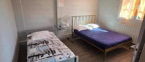 Una habitación pequeña con dos camas individuales, una ventana con persianas, un ventilador de pared y un interruptor de luz.