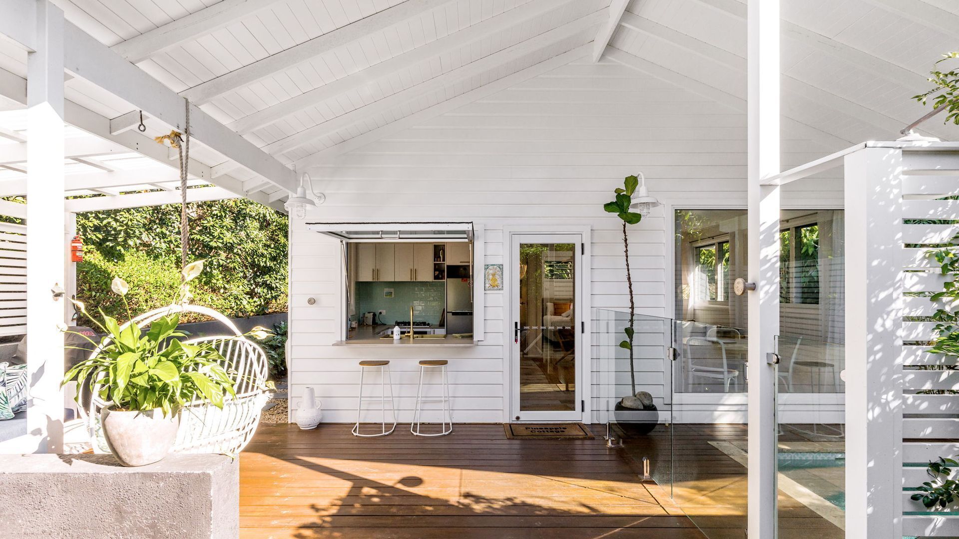 A covered outdoor area with a wooden deck, a kitchen with a window, and a door leading to a glass door.