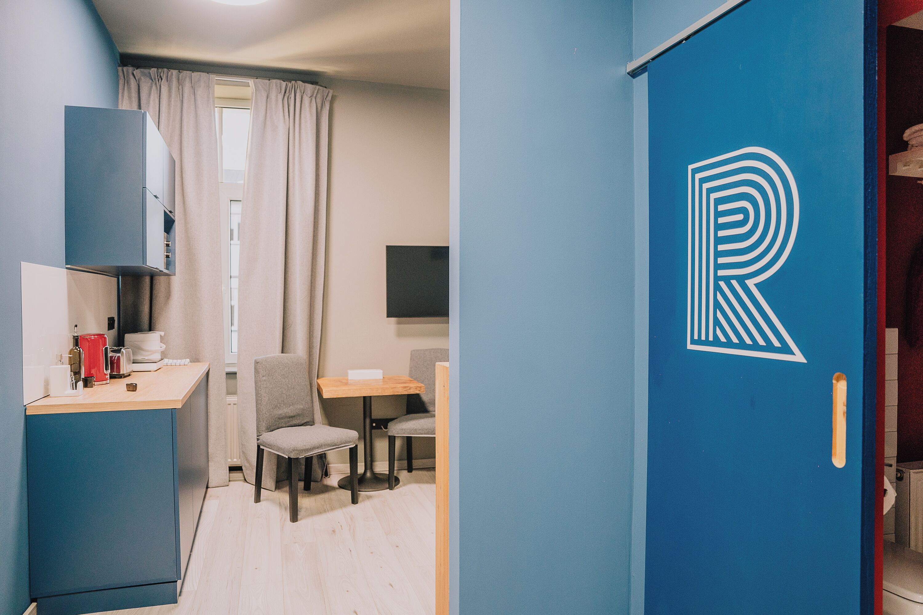 A compact room with a kitchenette, a dining area, and a door with a large letter 'R' on it.