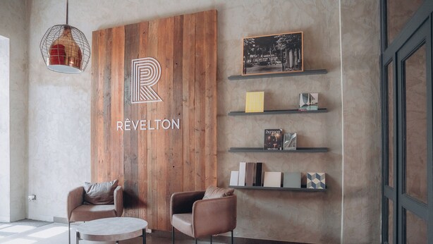 A modern interior with a wooden accent wall, a logo, two armchairs, a round table, and shelves with various items. - Revelton Ostrava (Ostrava)