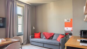 A modern hotel room with a grey sofa, red pillows, a wooden desk, a flat-screen TV, and a bed with white bedding. - Revelton Ostrava (Ostrava)