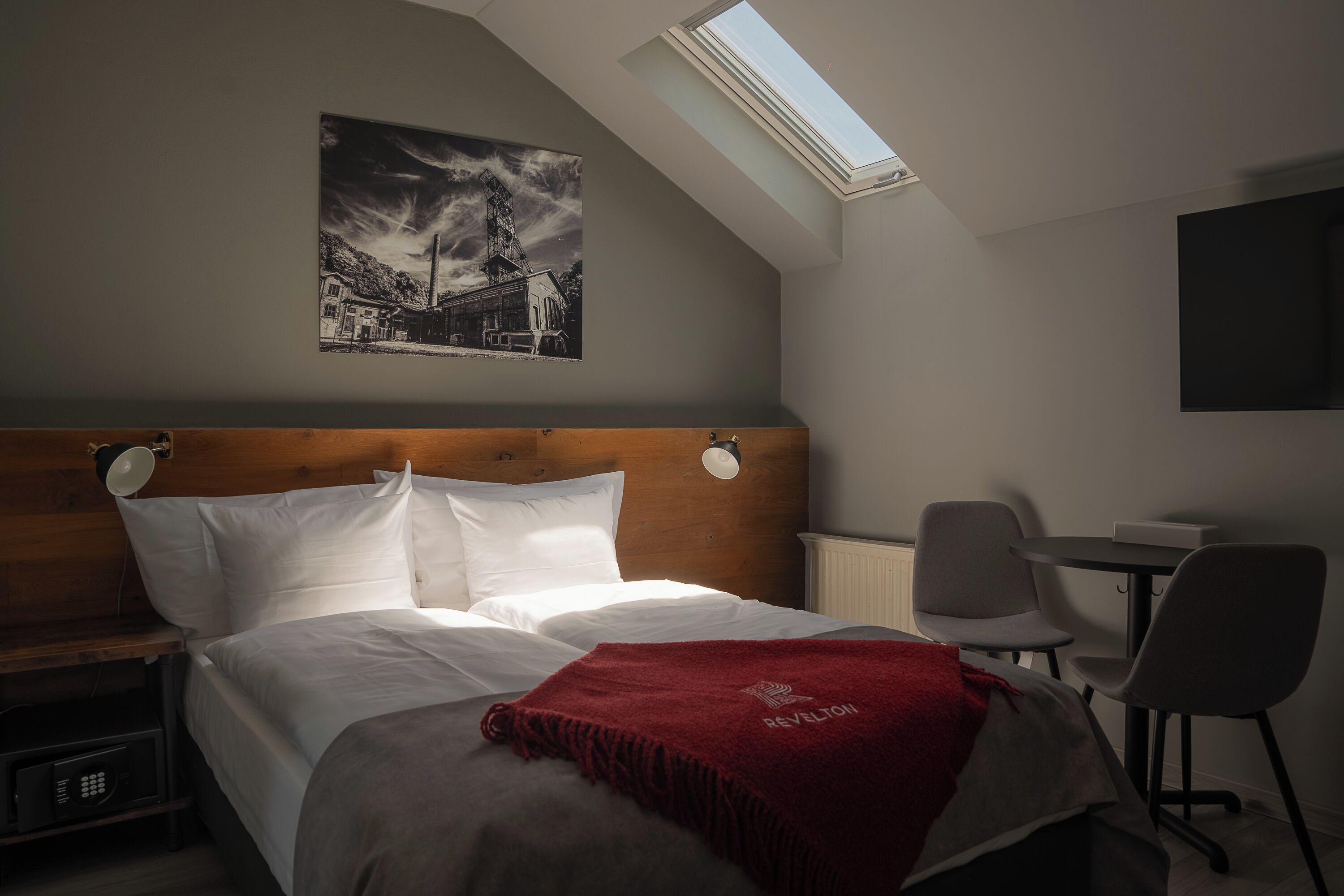 A bedroom with a skylight, a bed with white bedding, a red throw blanket, a wooden headboard, a bedside table with a lamp, and a chair.