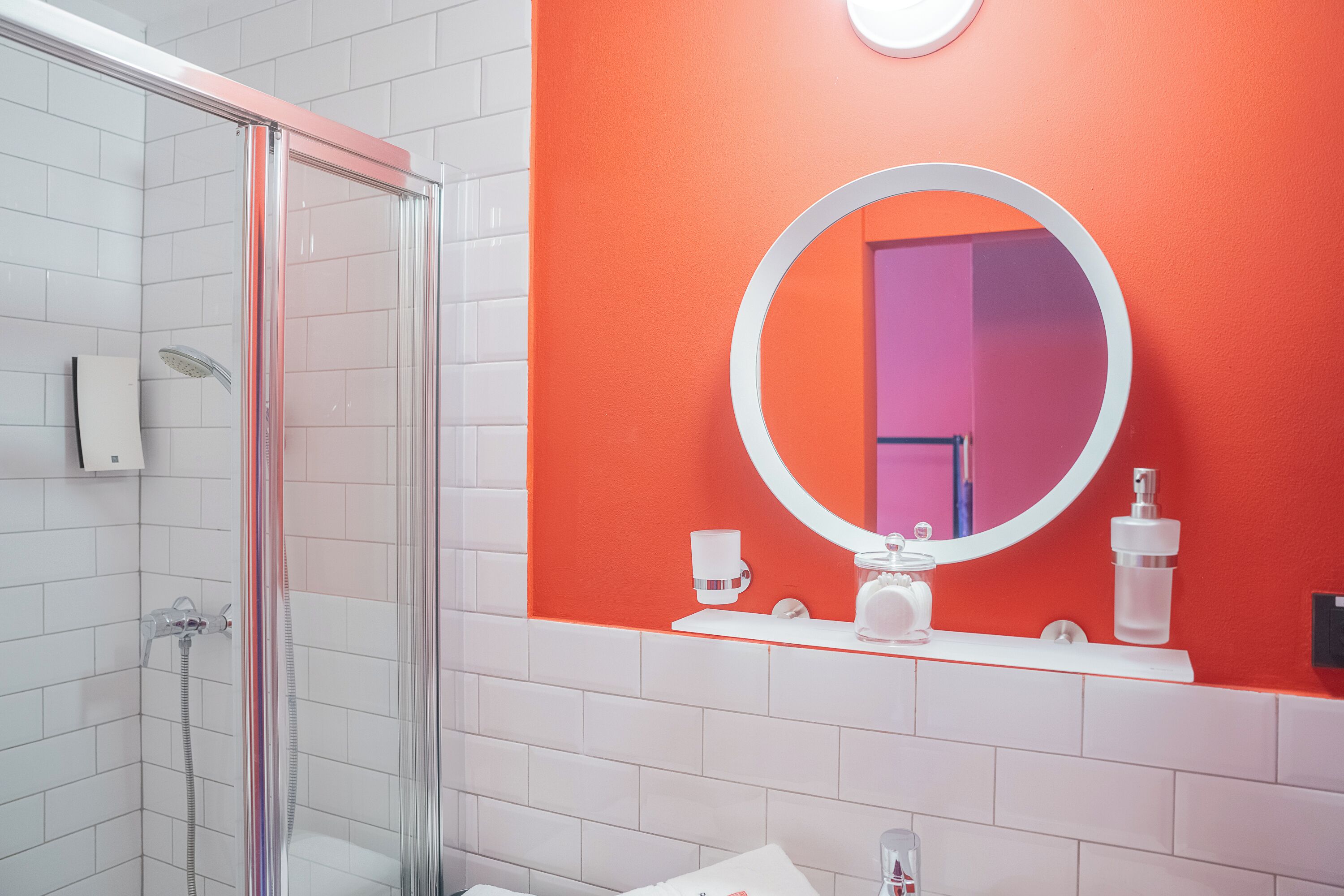 A bathroom with a glass-enclosed shower, a white sink, a round mirror, and a red accent wall.