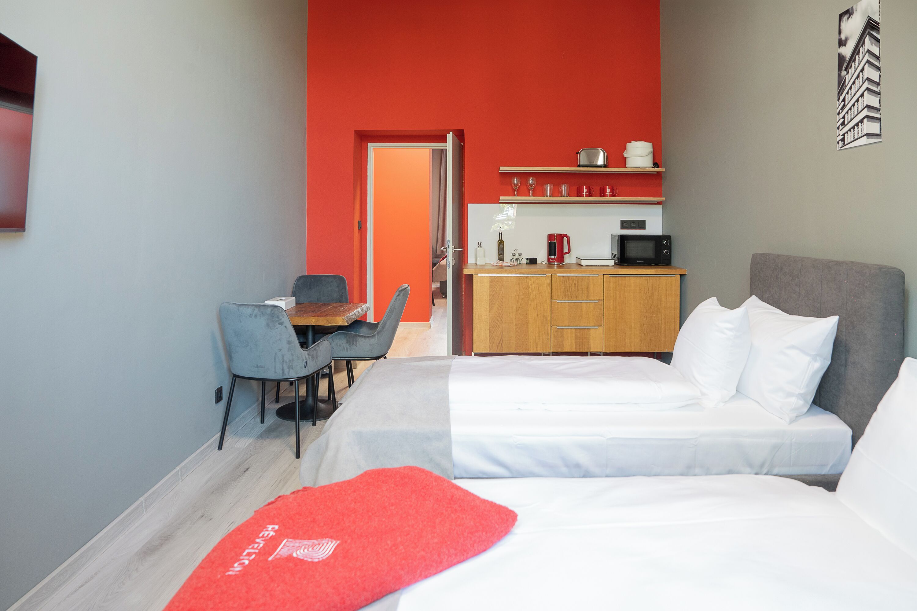 A hotel room with a bed, a desk with a chair, a kitchenette, and a red accent wall.