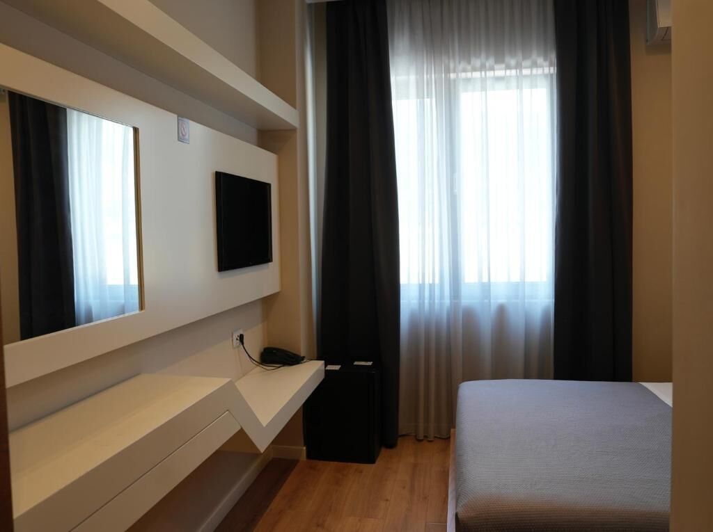 A hotel room with a bed, a TV mounted on the wall, a desk with a phone, and a window with curtains.