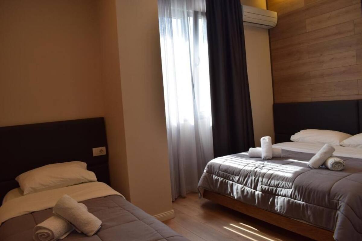 A hotel room with two beds, a window with curtains, a wooden headboard, and a wall-mounted air conditioning unit.