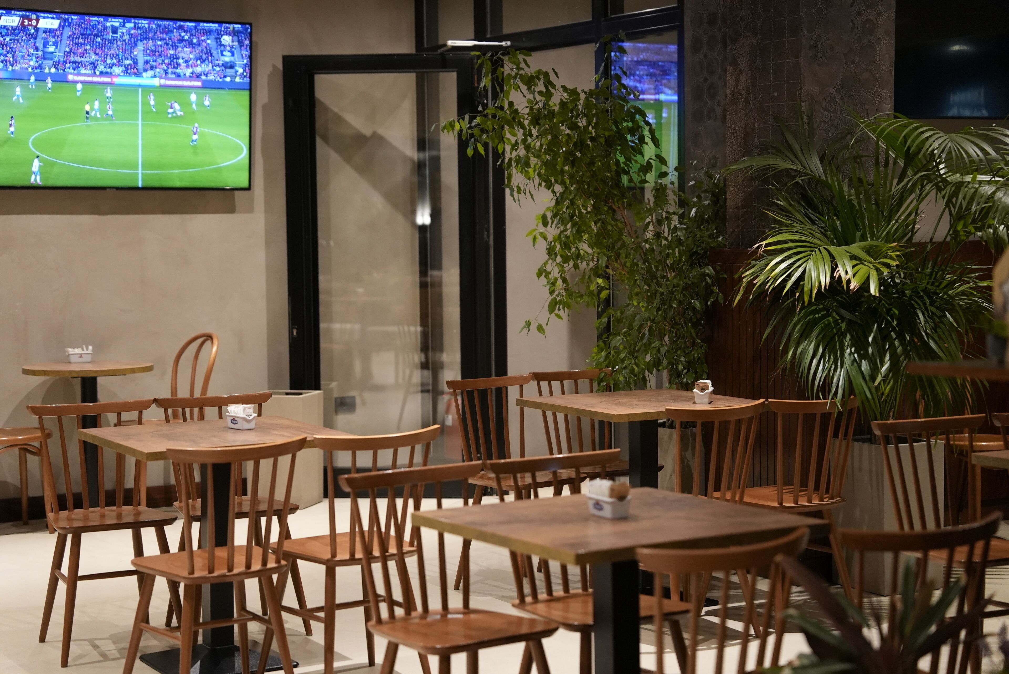 A restaurant with wooden tables and chairs, a television displaying a soccer match, and indoor plants.