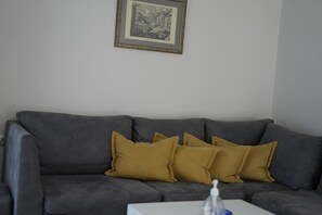 Un sofá gris con almohadones color mostaza, un cuadro en la pared y una mesa blanca con una botella.