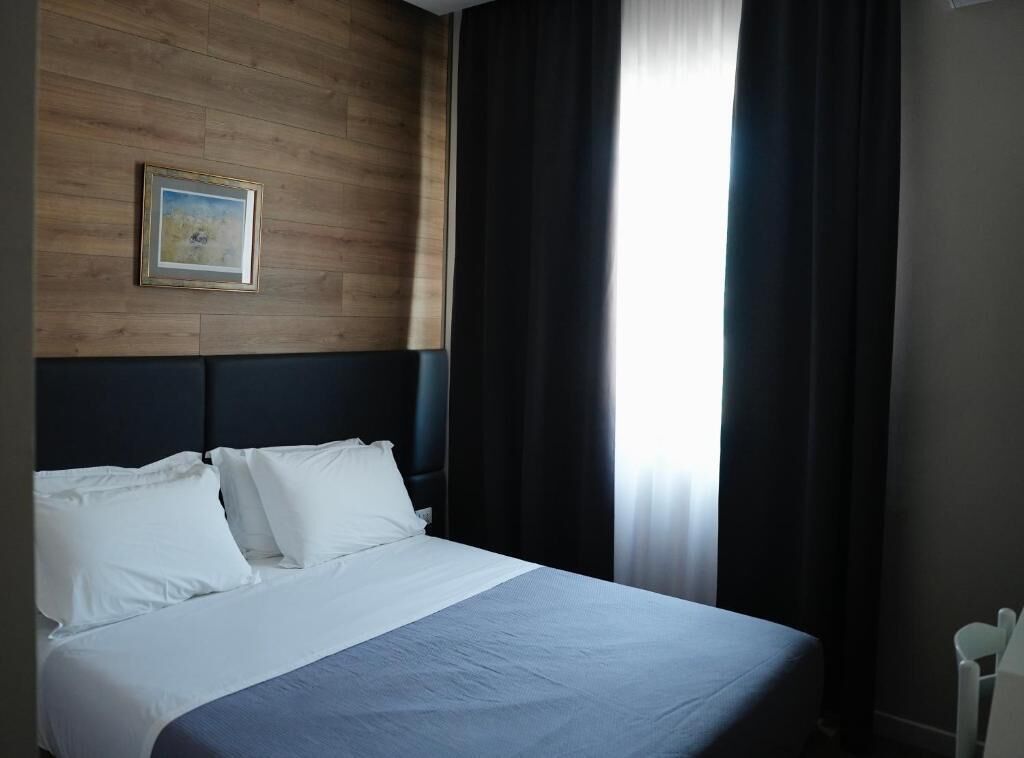 Habitación de hotel con cama, cabecera, un cuadro en la pared y una ventana con cortinas.