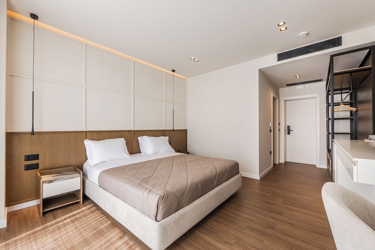 A modern hotel room with a large bed, bedside tables, a wardrobe, and a desk with a chair.