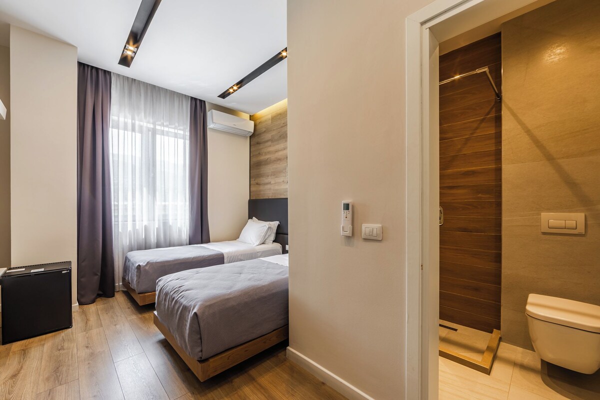 A hotel room with two beds, a small kitchenette, and a bathroom with a wooden floor.