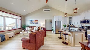Un espacio habitable amplio con un gran ventanal, un sillón de cuero marrón, un sofá y una cocina con refrigerador y microondas de acero inoxidable.