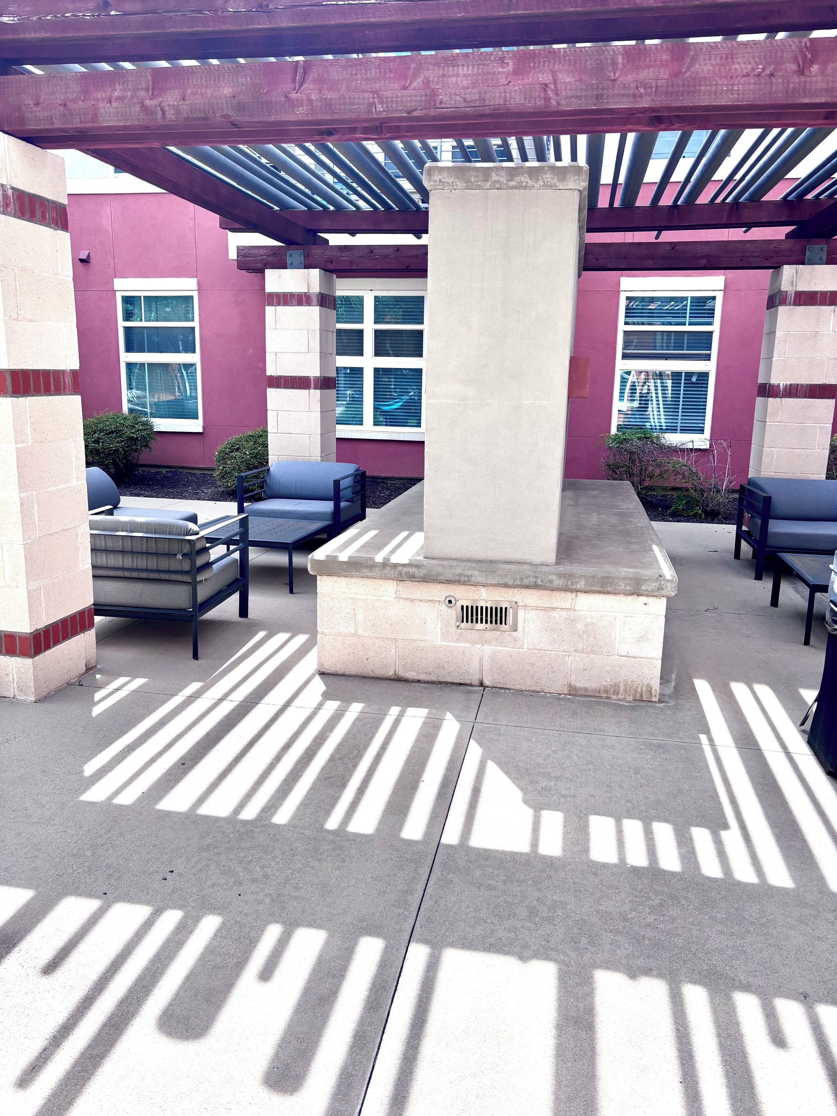 Un espacio al aire libre con bancos de hormigón y una estructura central de piedra, posiblemente una chimenea, bajo una pérgola de madera con vistas a un edificio con ventanas.