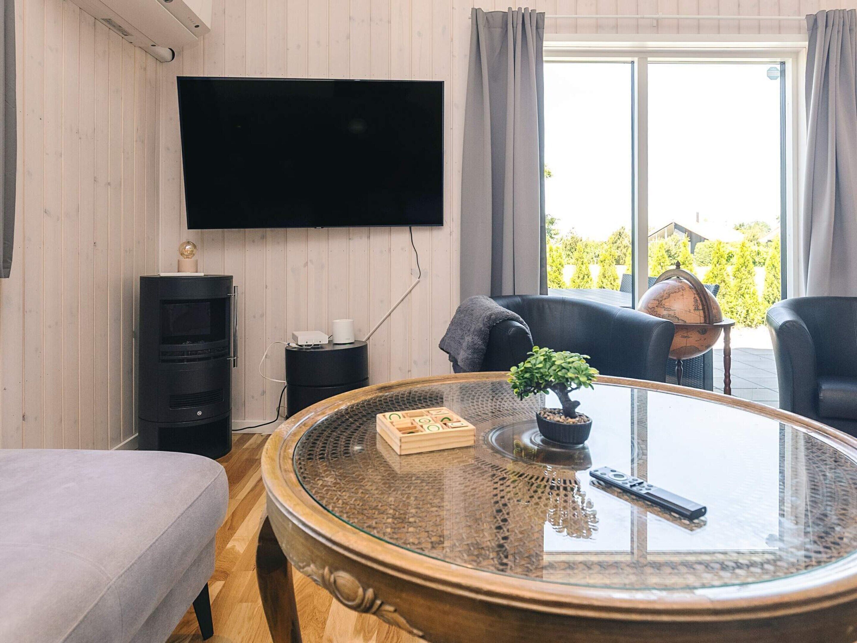 A modern living room with a large round glass coffee table, a flat-screen TV mounted on the wall, a black entertainment unit, and a view of greenery outside.