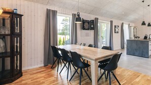 En rummelig spiseafdeling med et træbord og sorte stole, et moderne køkken i baggrunden og et stort vindue med gardiner.