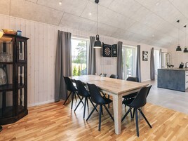En rummelig spiseafdeling med et træbord og sorte stole, et moderne køkken i baggrunden og et stort vindue med gardiner.