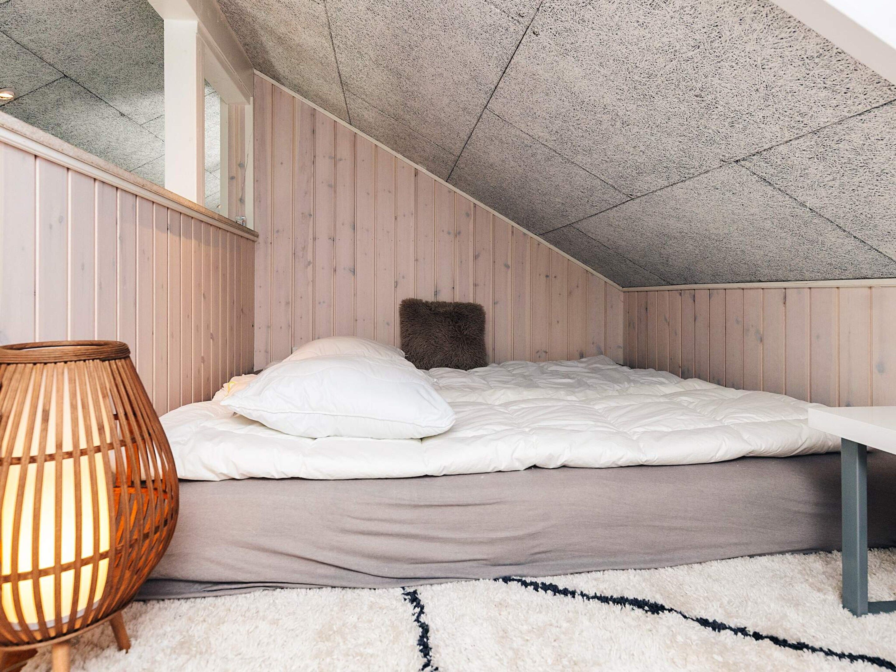 A bedroom with a sloped ceiling, wooden walls, a bed with white bedding, a fluffy pillow, a woven floor lamp, and a white rug.