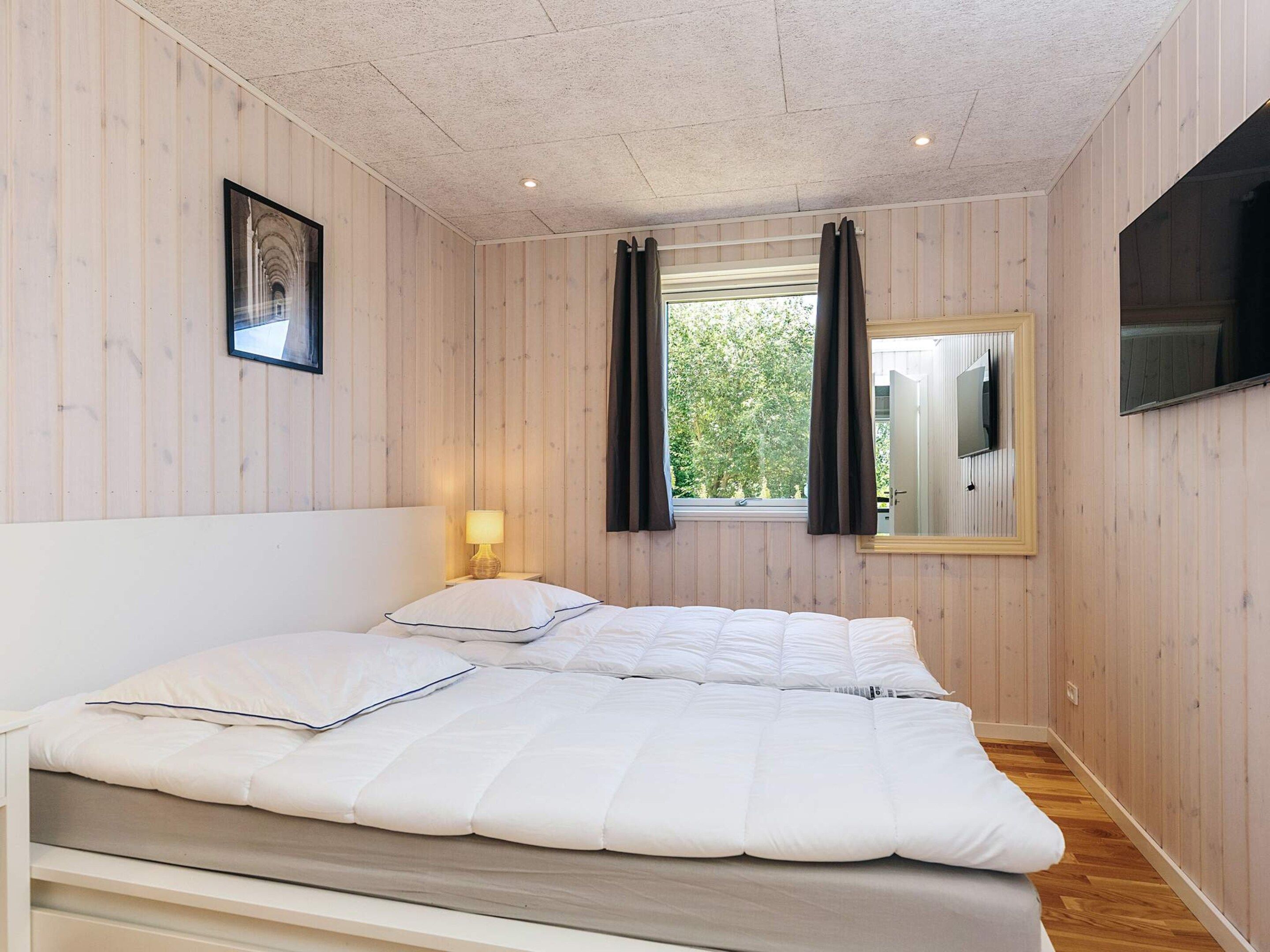 A bedroom with a bed, a bedside lamp, a window with curtains, and a flat-screen TV mounted on the wall.