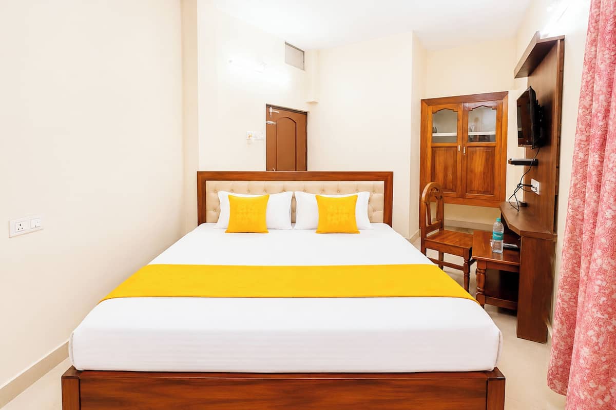 Habitación de hotel con una cama, una mesita de noche de madera, una silla, un escritorio, un televisor y una puerta.