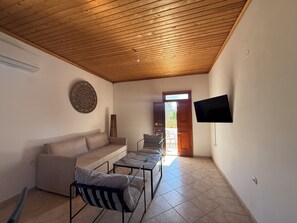 A living room with a wooden ceiling, a sofa, a chair, a TV, and a door leading outside. - Lambis Garden Hideaway (Lindos)
