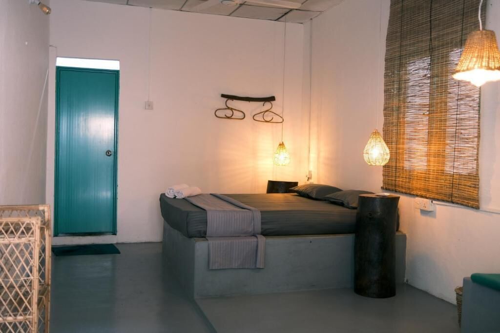 A single bed with a headboard, a bedside table, a lamp, a window with blinds, and a door.
