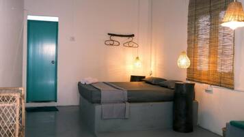 A single bed with a headboard, a bedside table, a lamp, a window with blinds, and a door.