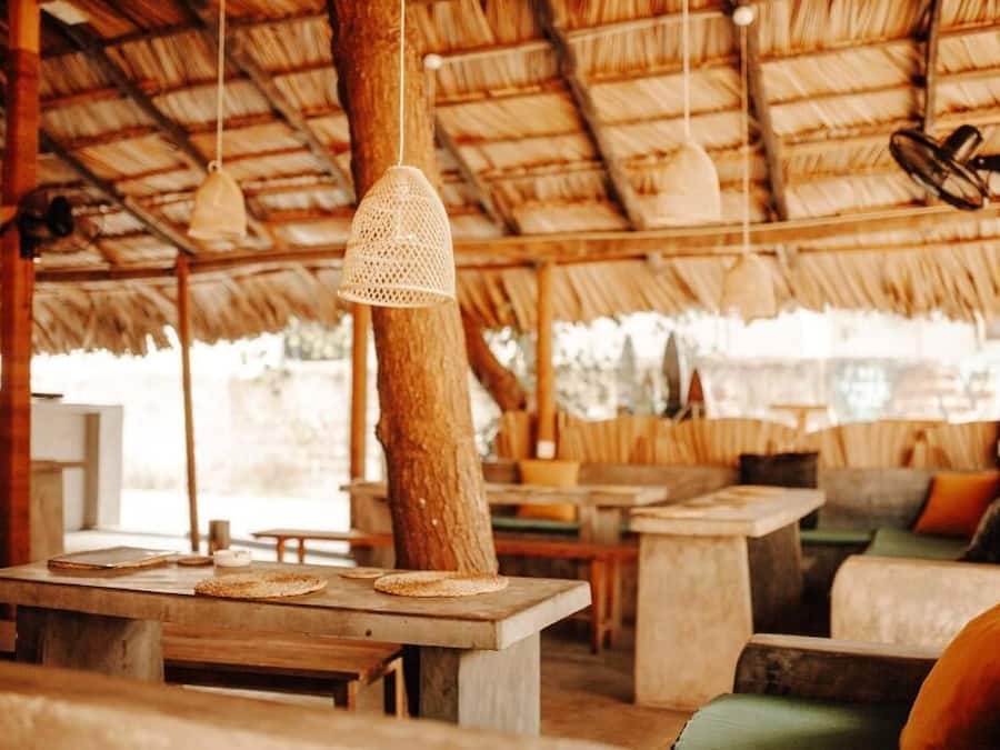 A rustic outdoor seating area with wooden tables and benches, a thatched roof, and hanging light fixtures.