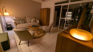 Ein Wohnzimmer mit einer Couch, Sesseln, einem Couchtisch, einer Lampe und einer Glastür, die auf einen Balkon führt.