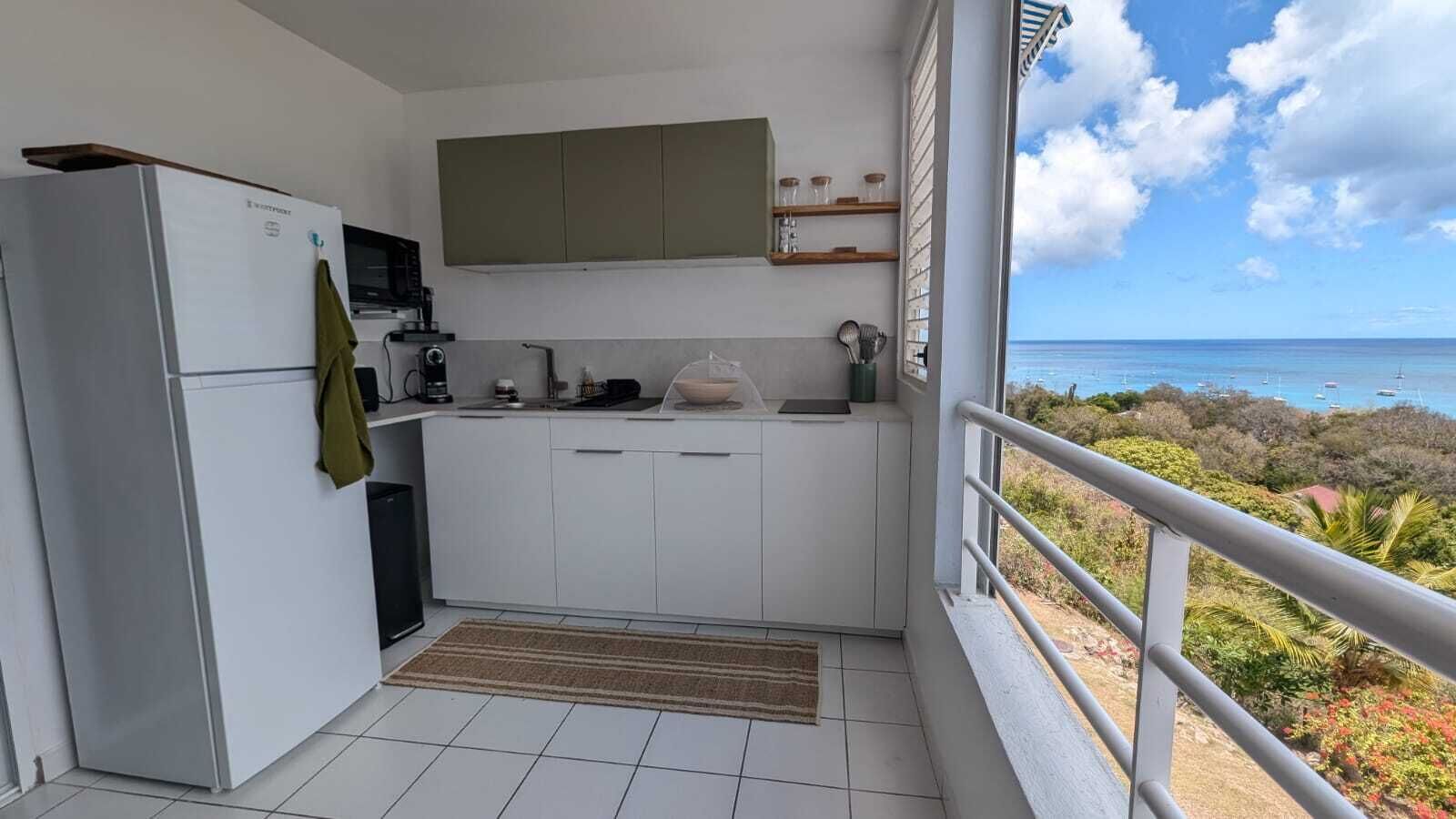 Uma cozinha compacta com geladeira, micro-ondas e cafeteira, uma pia pequena e uma janela com vista para o litoral.