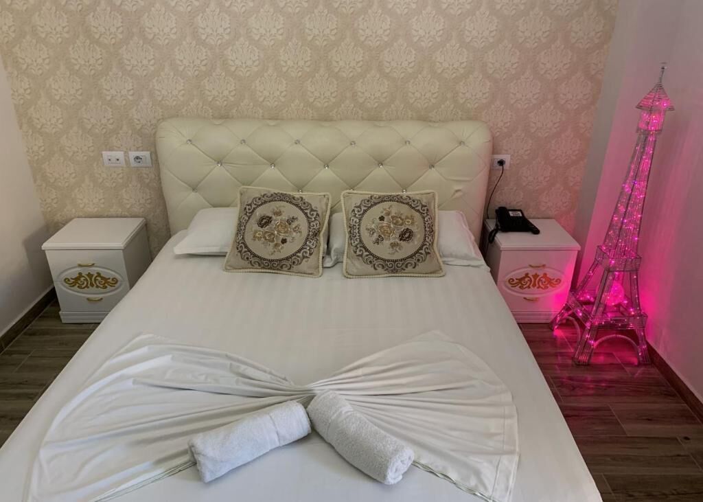 A bed with a tufted headboard, two decorative pillows, a folded towel, bedside tables with a phone and a lamp, and a pink Eiffel Tower decoration.