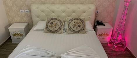 A bed with a tufted headboard, two decorative pillows, a folded towel, bedside tables with a phone and a lamp, and a pink Eiffel Tower decoration.