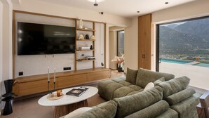 A modern living room with a flat-screen TV, a sofa, a coffee table, and a view of a mountainous landscape. - Azur Blue Luxury Villa (Mylopotamos)