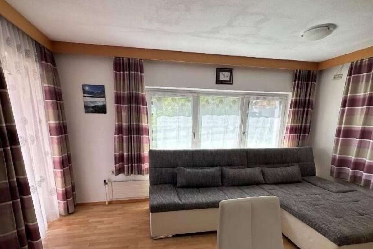 A room with a sofa bed, a chair, a table, and curtains.