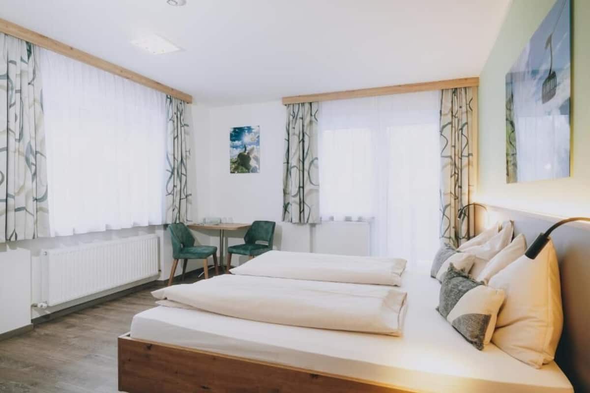 A hotel room with a bed, a desk, two chairs, and large windows with curtains.