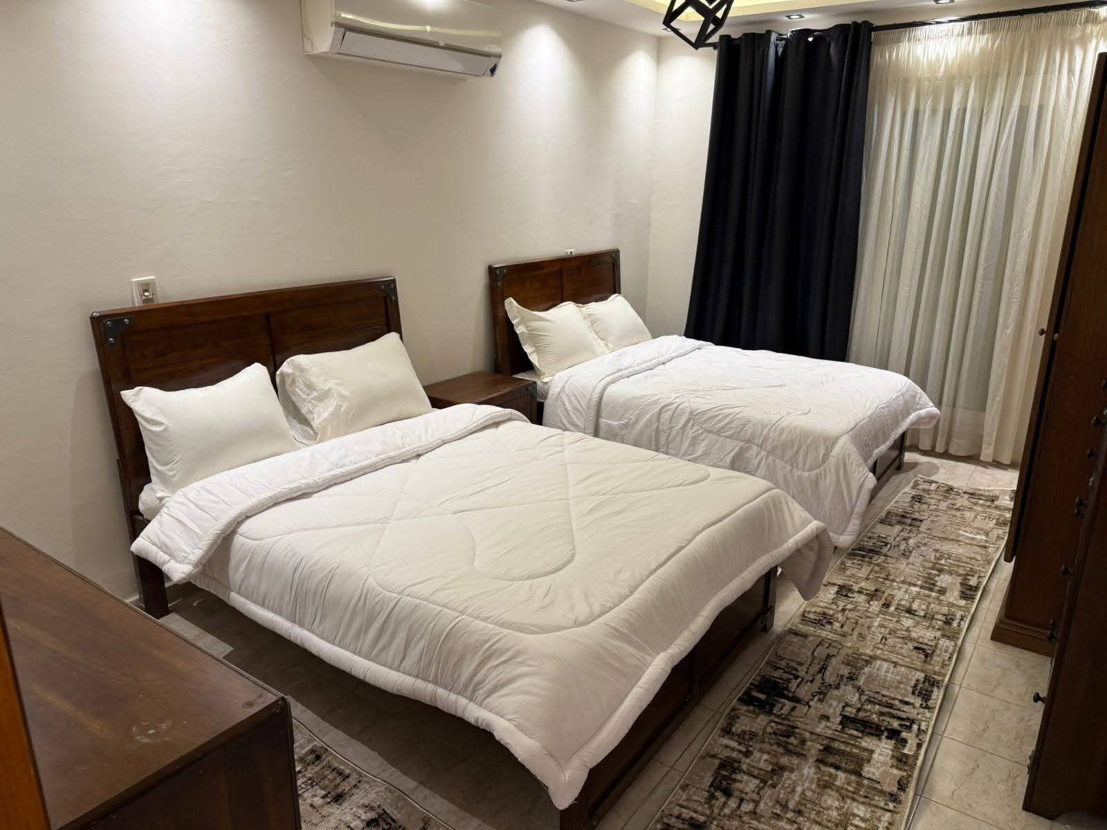 A double room with two beds, a wooden headboard, a bedside table, a rug, and a window with curtains.