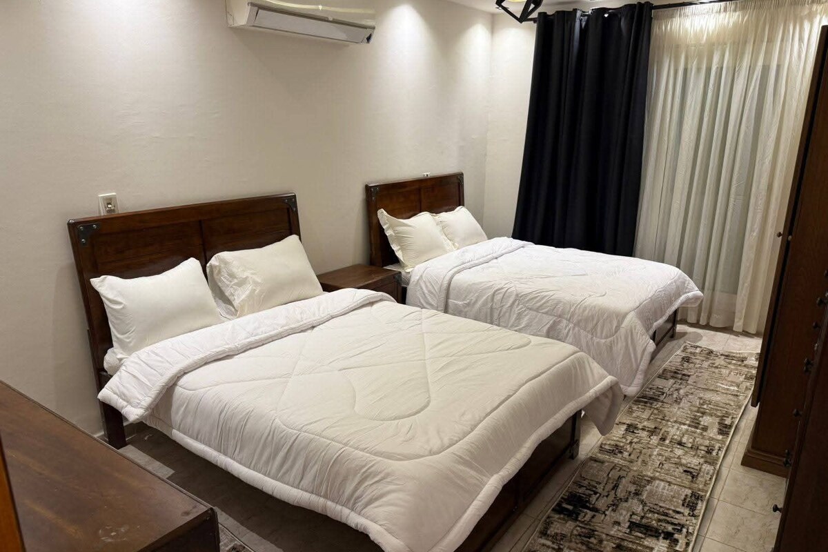 A double room with two beds, a wooden headboard, a bedside table, a rug, and a window with curtains.