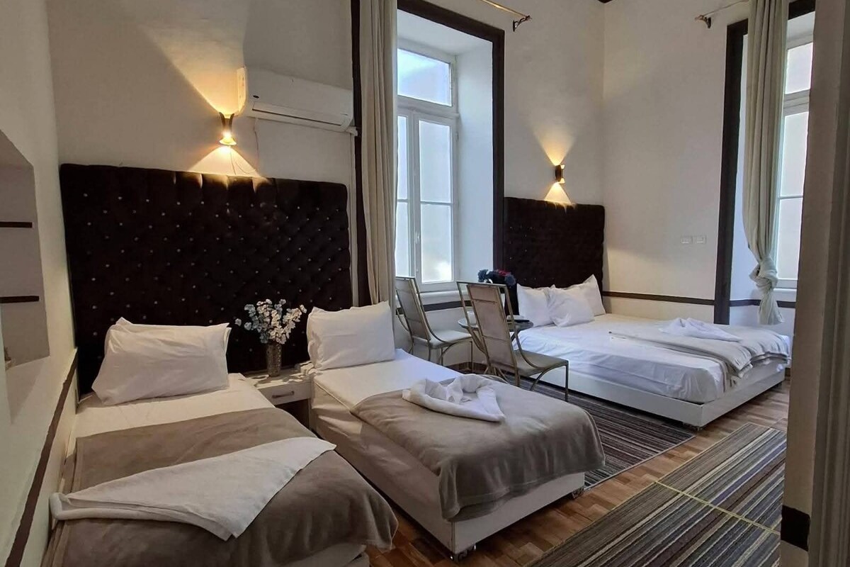 A hotel room with two beds, a chair, a small table with a vase of flowers, and a window with curtains.
